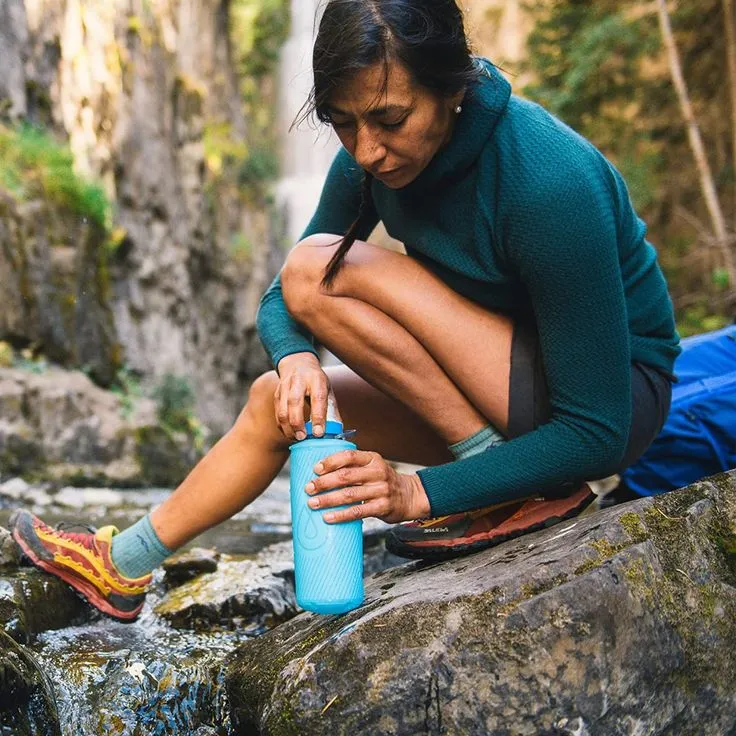 A backpacker refilling a collapsible water bottle from a stream—highlighting How Much Water to Bring When Backpacking.
