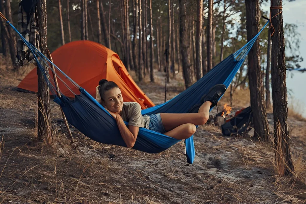 A camper relaxes in one of the best hammocks, strung between trees at a forest campsite near a glowing fire and orange tent.
