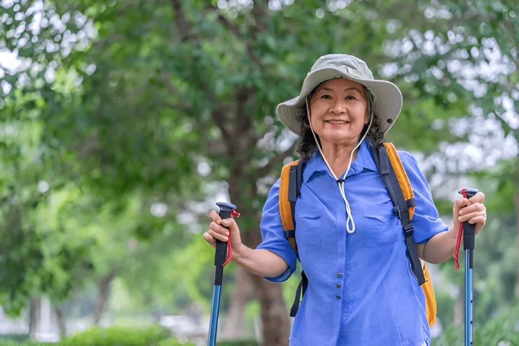 portrait cheerful senior woman wears hat,with backpack and trekking poles standing in nature,elderly asian female happy in hiking trip